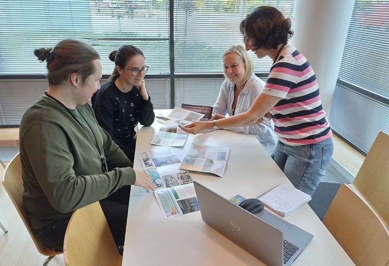 Salon kaupungin maahanmuuttajapalveluiden neljä työntekijää pöydän ääressä kabinetti Tuomessa Salon kaupungintalolla. Pöydällä on esitteitä. Yksi henkilöistä seisoo ja pitää esitettä kädessään. Muut kolme henkilöä istuvat ja hymyilevät.