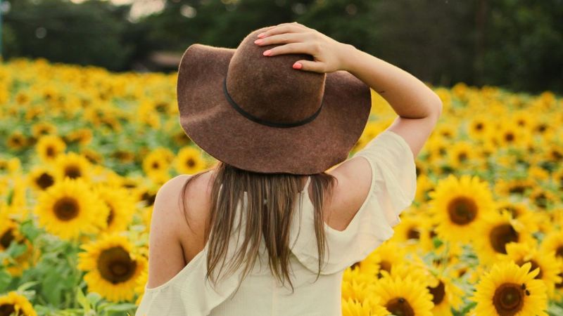 Nuori pitkähiuksinen nainen katselee auringonkukkapeltoa. Hän pitää oikealla kädellään kiinni päässään olevasta ruskeasta leveälierisestä kesähatusta. Nainen on selin katsojaan. 