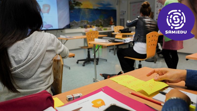 Kuvassa henkilö kulkee opetustilassa. Henkilöitä istuu työpöytien ääressä tekemässä tehtäviä, joissa on suomen kielen sanoja. Näytöllä on sohvan kuva ja teksti sohva.