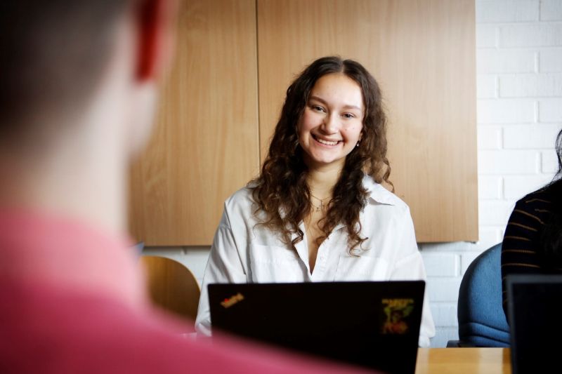Nuori nainen istuu kannettavan tietokoneen ääressä ja katsoo hymyillen edessään istuvaa henkilöä.