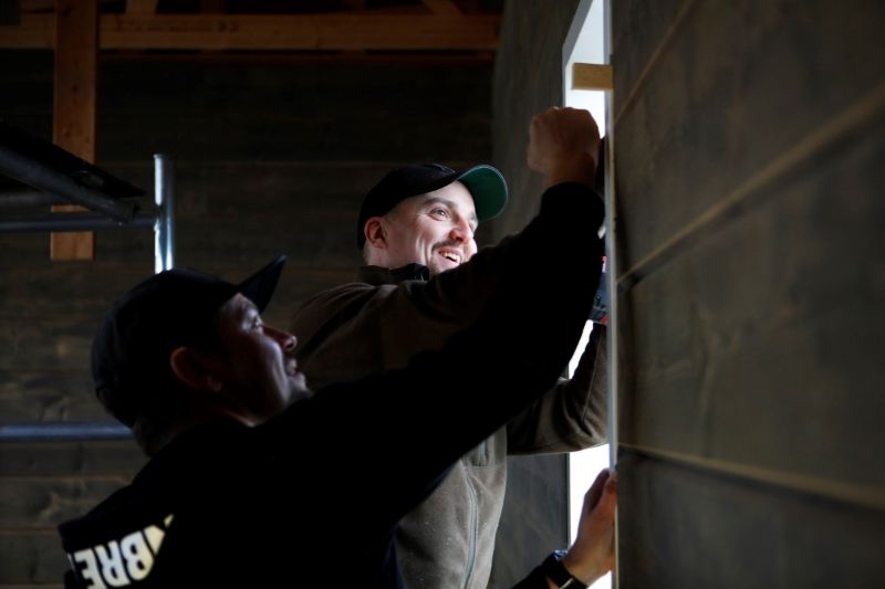 Rakentajat asentavat yhdessä jotain keskeneräisen ikkunan karmiin rakennuksen sisällä.