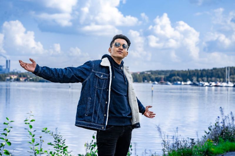 Henkilö seisoo aurinkolasit päässä kädet levällään järvimaiseman edessä. Taustalla järven takana näkyy metsää, satama ja tehdas.