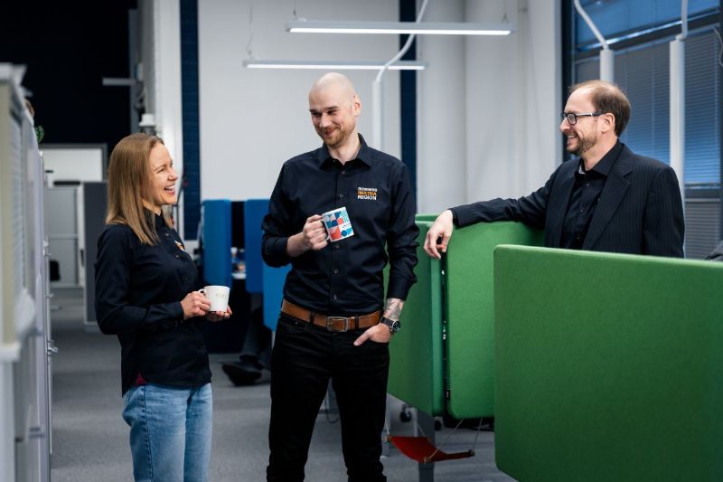 Kolme henkilöä seisovat toimistoympäristössä kahvikupit kädessään.