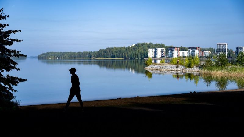 Henkilön siluetti rannalla kävelemässä. Taustalla järvimaisema ja kauempana kerrostaloalue.