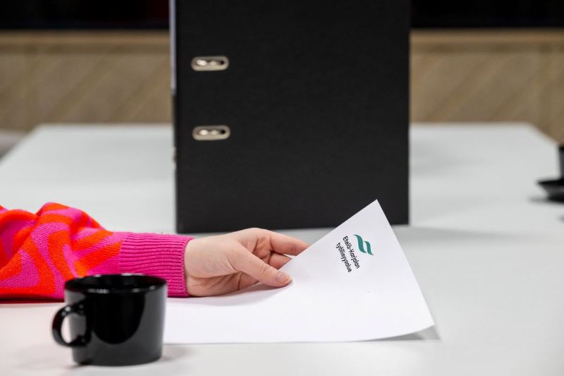 Käsi pitelee paperia, jossa Etelä-Karjalan työllisyysalueen logo. Etualalla kahvikuppi ja taustalla mappi.