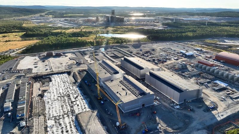 Ilmakuva suuressa teollisuusalueesta, jossa on rakenteilla useita suuria tehdashalleja ja nosturi työmaalla. Etualalla näkyy rakennusalueita, työmaakoneita ja tilapäisiä rakennuksia. Taustalla levittäytyy vihreä metsäalue ja kauempana näkyy olemassa olevia teollisuusrakennuksia sekä vesialueita, joiden pinnasta heijastuu auringonvalo.