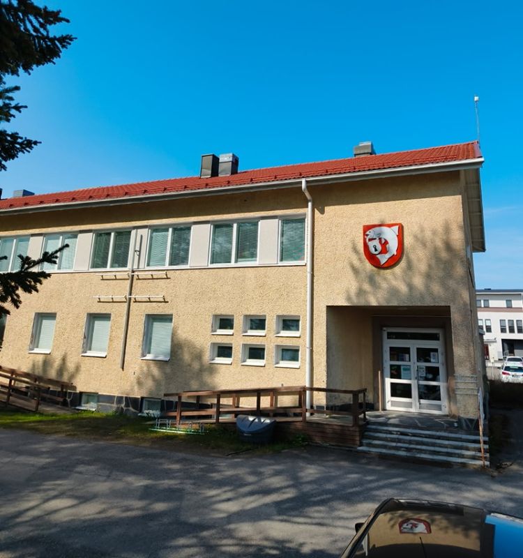 Vaalea kaksikerroksinen rakennus, jonka julkisivussa on punakatto ja seinässä vaakuna. Rakennuksen edessä on portaat ja luiska sisäänkäynnille, ja taustalla näkyy sininen taivas ja muita rakennuksia.