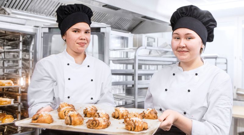 Kaksi opiskelija esittelevät leipomiaan pullia tarjottimella.