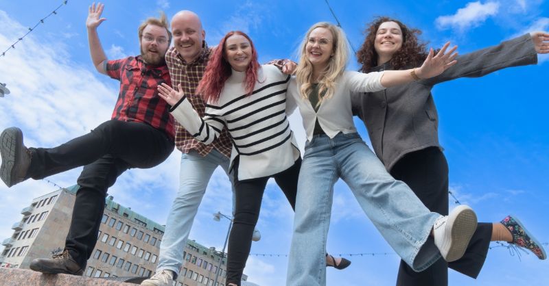 Viisi henkilöä vilkuttavat ja hymyilevät kameralle sinistä taivasta vasten.