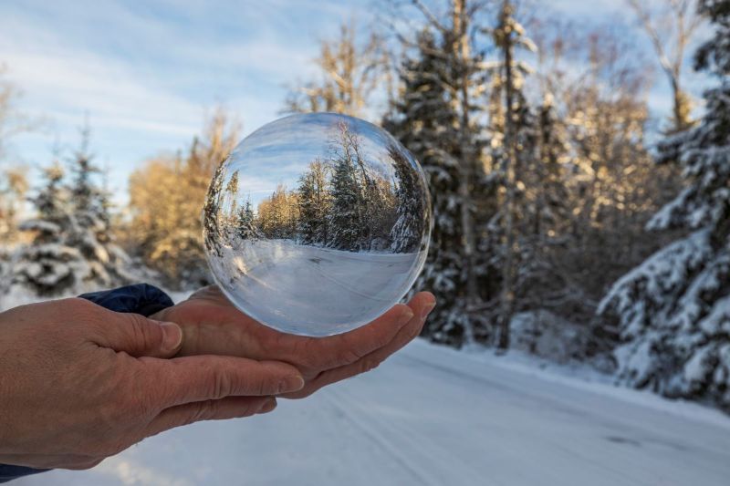 Kuvituskuva. Kuvassa kädellä olevasta lasipallosta heijastuu kuva taustalla olevasta talvisesta maisemasta.