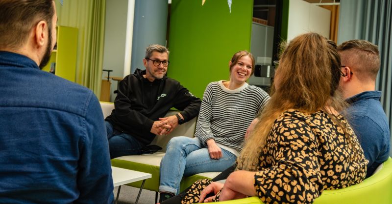 Henkilöt keskustelevat ja nauravat sohvalla istuen.