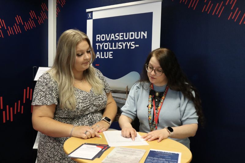 Kaksi henkilöä seisoo pienen pyöreän pöydän ääressä tutkimassa paperilomaketta. Taustalla on tummansininen seinä ja rullajuliste, jossa lukee "Rovaseudun työllisyysalue".