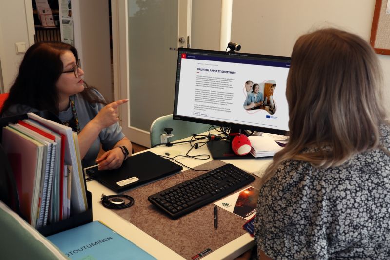 Kaksi henkilöä istuu toimistopöydän eri puolilla. Pöydällä on näppäimistö, läppäri ja eri värisiä papereita. Henkilöt ovat keskittyneet katsomaan verkkosivua, joka on avattu erilliselle tietokonenäytölle.