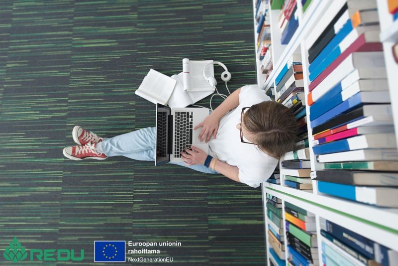 Henkilö istuu kirjaston lattialla kirjahyllyn edessä ja työskentelee kannettavalla tietokoneella kirjojen ja muistiinpanojen ympäröimänä.