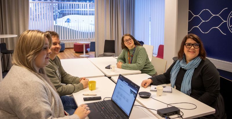 Työllisyyspalveluiden kokous neuvottelutilassa: neljä henkilöä istuu pöydän ääressä, pöydällä on kannettava tietokone, muistivihkoja ja kahvikuppeja.