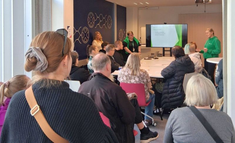 Noin 15 hengen ryhmä on kokoontunut kokoustilaan kuuntelemaan esittelyä. Ryhmä seuraa tiiviisi esitystä, jota pitävät REDUn työntekijät suurta televisioruutua hyödyntäen.