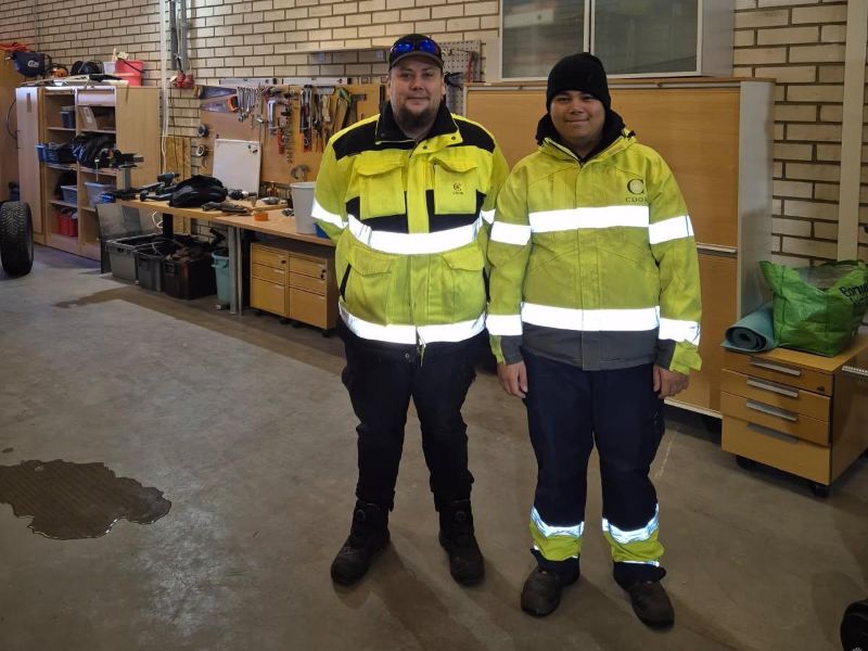 Kaksi Luovin opiskelijaa seisovat vierekkäin ja katsovat hymyillen kameraan. Heillä on päällään keltaiset työtakit. Taustalla on erilaisia työkaluja.