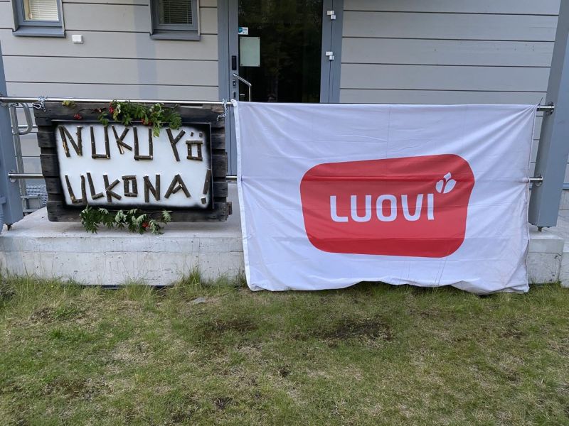 Terassin kaiteeseen kiinnitetty suuri valkoinen liina, jossa on Luovin punainen logo. Sen viereen on kiinnitetty luonnonmateriaaleista valmistettu kyltti, jossa on teksti "nuku yö ulkona".