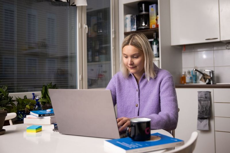Laventelin väriseen neulepuseroon pukeutunut, vaaleahiuksinen nuori henkilö istuu keittiönpöydän ääressä käyttämässä kannettavaa tietokonetta. Valkoisella pöydällä on tietokoneen lisäksi kahvimuki ja joitain kirjoja, taustalla on viherkasveja ikkunalla ja valkoisia keittiön kaappeja.