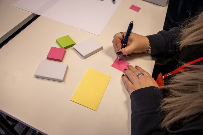 Henkilö kirjoittaa vaaleanpunaiselle post it -lapulle. Pöydällä on myös keltaisia, valkoisia ja vihreitä lappuja tyhjänä.