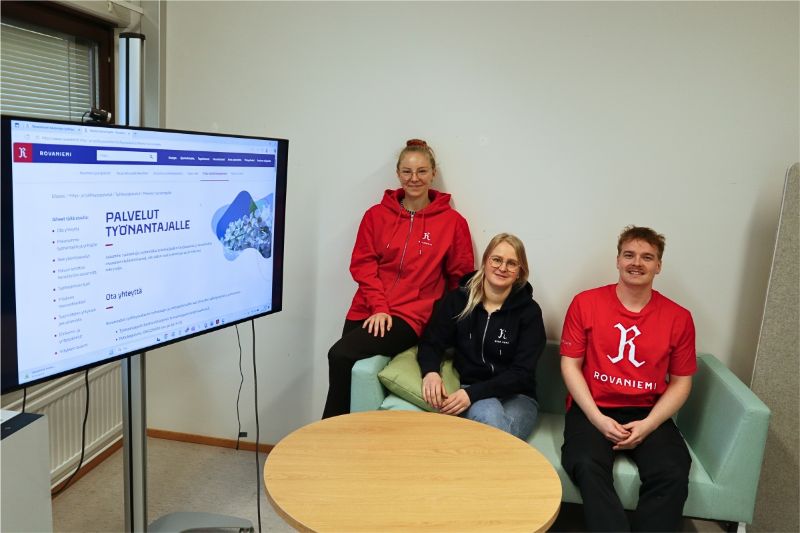 Yritysasiantuntijat Amanda Suokas, Miia Nurmela ja Juho Poikajärvi istuvat turkoosinvihreällä sohvalla ja katsovat hymyillen kameraan. Heidän vasemmalla puolellaan on iso tv-ruutu, jolla näkyy Rovaniemen kaupungin työnantaja- ja yrityspalvelujen verkkosivut.