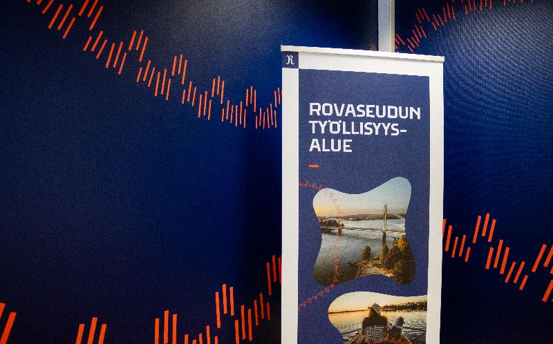 Rovaseudun työllisyysaluetta kuvaava rullajuliste. Taustalla on tummansininen seinä, johon on maalattu punaista, revontulia kuvastavaa symbolinauhaa.