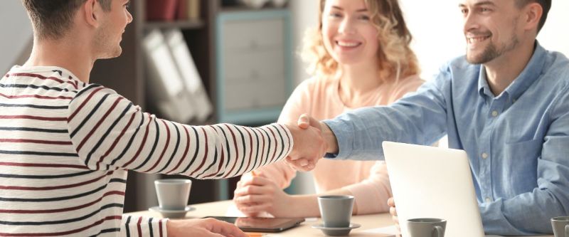 Kolme ihmistä pöydän äärellä, kaksi kättelevät. 