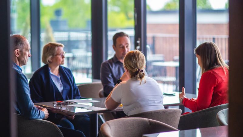 Kokoustilanne. Viisi henkilöä istuu pöydän ääressä. Pöydällä on papereita. Taustalla isot ikkunat.