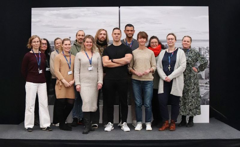 13 henkilön ryhmäkuva. Henkilöt seisovat lähekkäin pienellä korokkeella. Taustalla ilmakuva Tampereen keskustasta. Group photo of 13 people. The individuals are standing close together on a small platform. In the background, an aerial view of downtown Tampere.