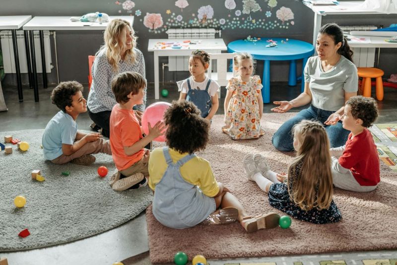 Ryhmä päiväkodin lapsia leikkimässä lattialla kahden ohjaajan kanssa. Yhdellä lapsista on käsissään iso ilmapallo. Huoneessa  pehmeitä mattoja, lapsille sopivia huonekaluja ja erilaisia leikkivälineitä.<br/>A group of preschool children playing on the floor with two instructors. One of the children is holding a large balloon. The room has soft rugs, child-friendly furniture, and a variety of toys.